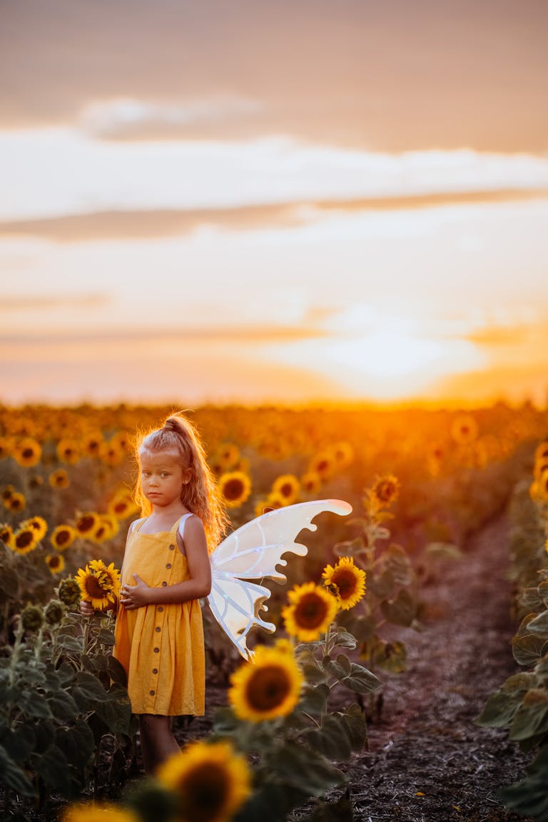 A young girl in a sunflower field at sunset, wearing fairy wings and a yellow dress.