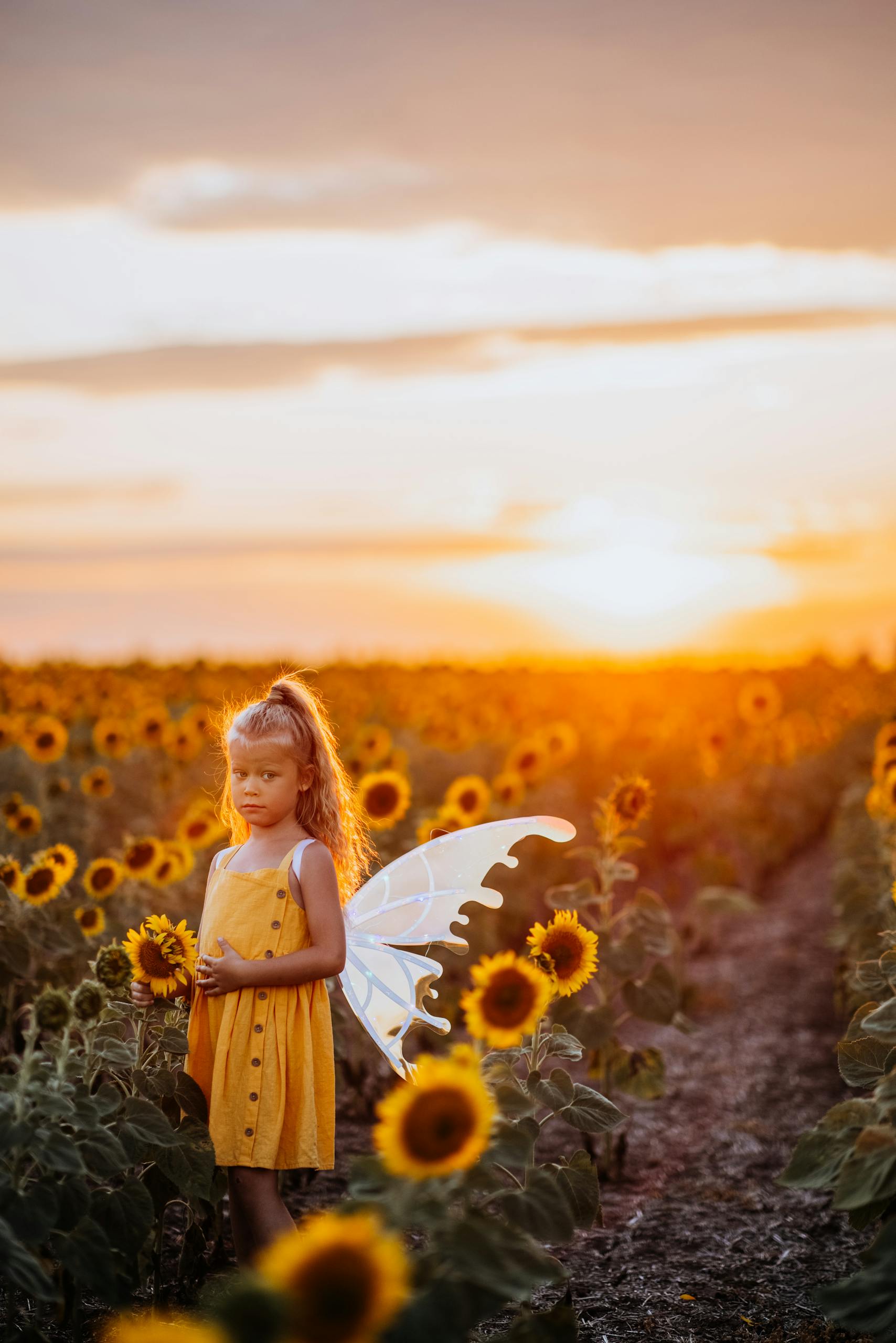 A young girl in a sunflower field at sunset, wearing fairy wings and a yellow dress.