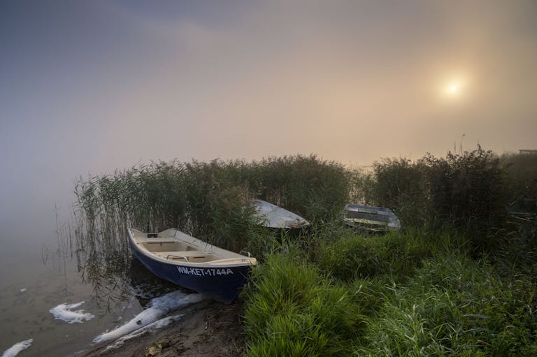 Serene misty sunrise over a lake in Poland with boats and lush greenery.