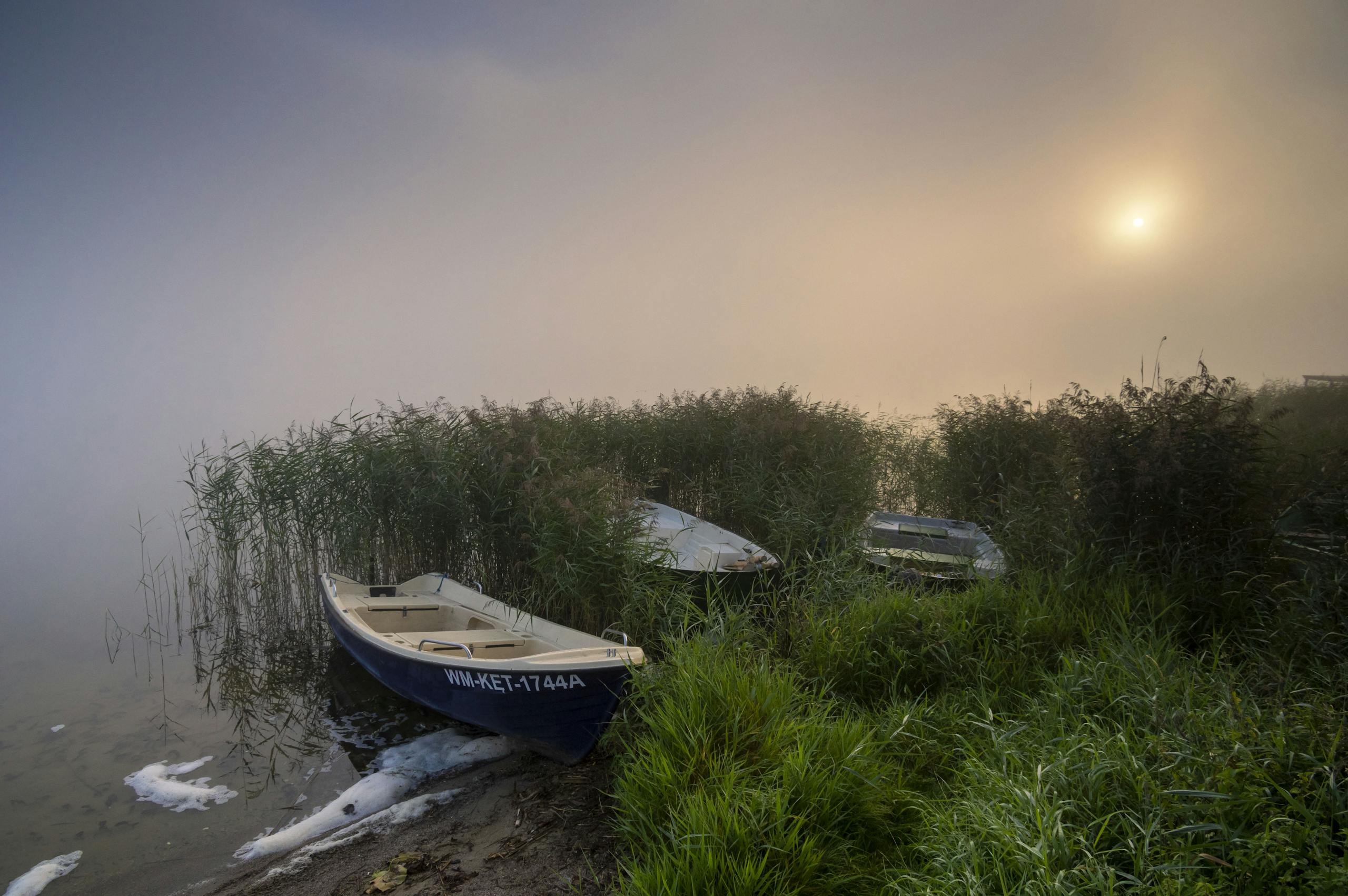 Serene misty sunrise over a lake in Poland with boats and lush greenery.